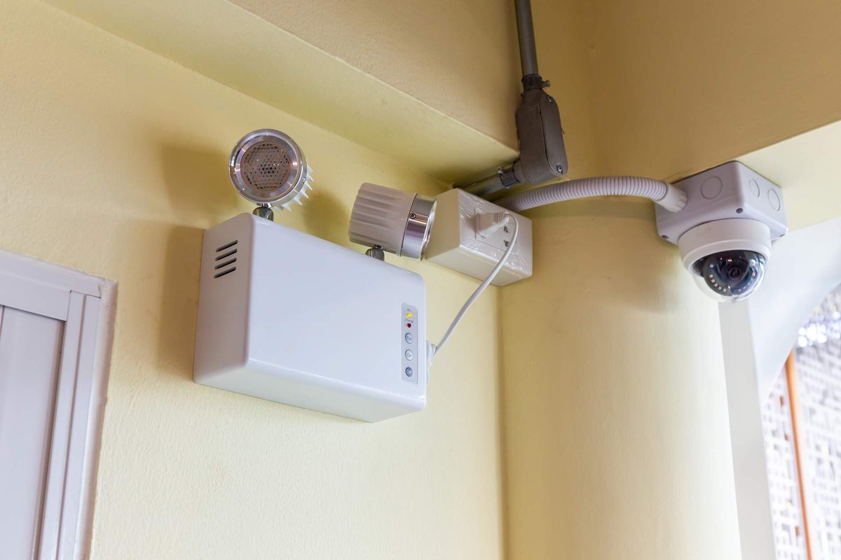 A twin-headed emergency light unit and a dome security camera mounted where a wall meets the ceiling, wired through grey conduit