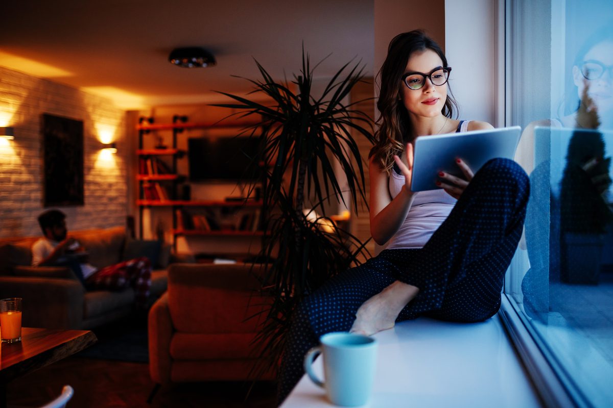 A woman reading a tablet on a window seat in the evening, with wall lights glowing on a brick wall and lit bookshelves in the lounge behind her