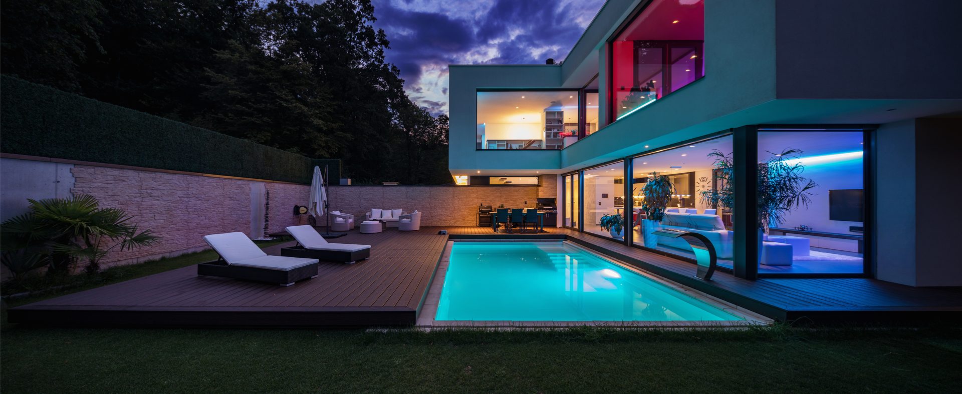 A modern flat-roofed house at dusk with its rooms lit, a glowing blue swimming pool, a timber deck with two sun loungers and a lawn in front