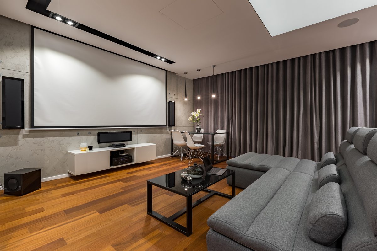 A home theatre lounge with a large projector screen, speakers on a low cabinet, a grey corner sofa, timber floors and a line of ceiling downlights