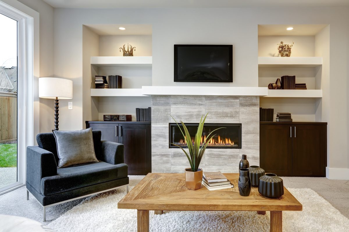 A bright lounge with a gas fire set in a stone surround, a television above it and built-in shelves on each side lit by small downlights
